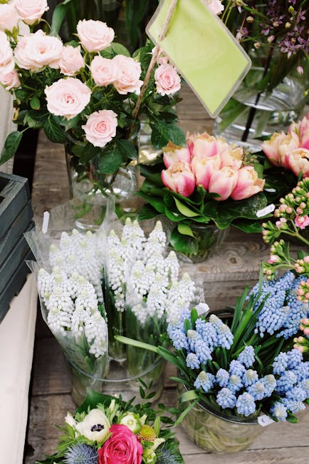A display of fresh floral arrangements featuring light pink roses in a clear glass vase, complemented by pink and cream tulips arranged in a round bouquet. Surrounding these are clusters of white grap