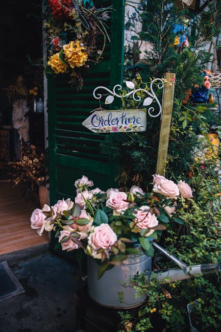 A charming outdoor floral display featuring a large bouquet of pale pink roses with lush green foliage placed inside a vintage-style silver watering can, situated on the ground amidst various flowerin