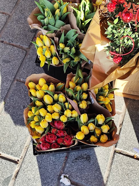 Several bouquets of fresh flowers displayed outdoors on a paved surface, including yellow tulips with green stems and leaves arranged in brown wrapping paper, red roses within a brown paper wrap, and